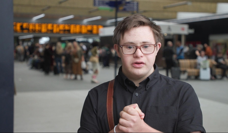 A man stands in a black button-up shirt in a train station