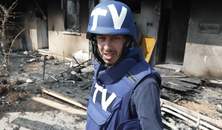 Secunder Kermani walks through rubble in a body armour vest and helmet, both of which have 'TV' written on them in tape