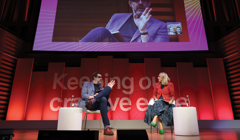 Richard Osman and Marina Hyde talk onstage at the RTS London Convention, beneath a large screen displaying a live feed of Osman