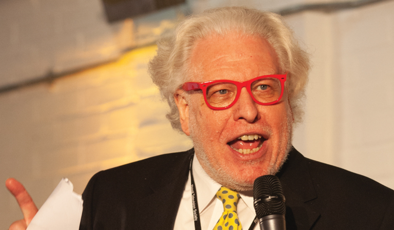 Simon Albury speaking publicly, wearing distinctive red glasses