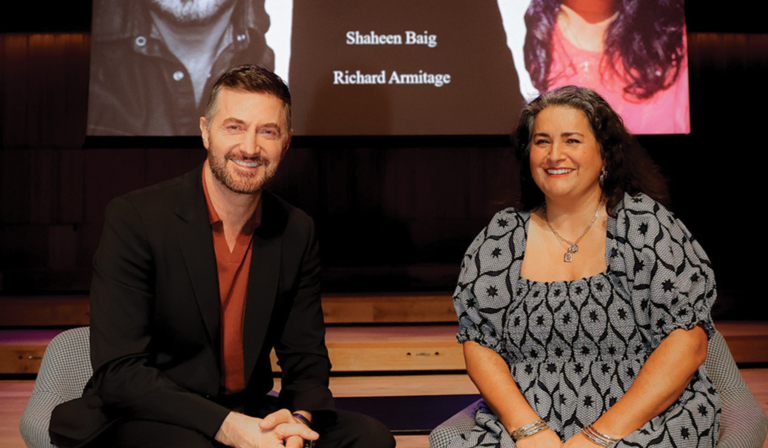 Shaheen Baig and Richard Armitage smile into the camera, sat down in front of a projector screen showing both their names