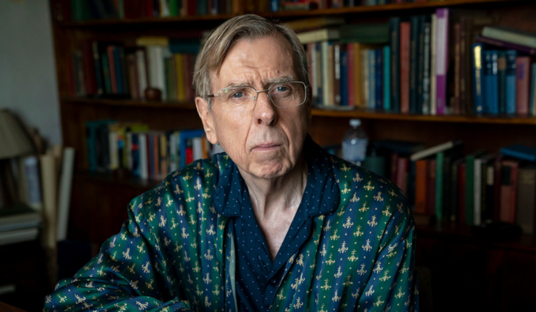 Timothy Spall, a man in his mid sixties, sits in front of a large fully packed bookcase, in his blue polkadotted pyjamas and a green and navy striped silk dressing gown. He wears rectangular glasses.