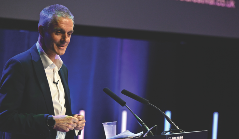 Tim Davie, who has short white hair and is in his mid-fifties, stands at a podium, smiling slightly