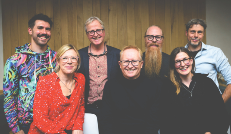 The seven people stand indoors in front of wood panelling, smiling to the camera