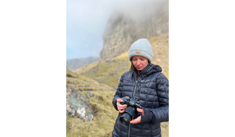 Ellie Green holds a camera while standing on a grassy and rocky mountain