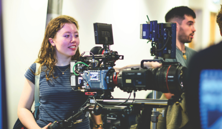 A young woman in a stripy top operates a large, industry-standard video camera