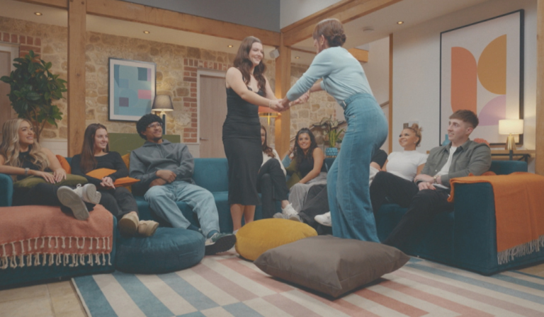 Two woman join hands, facing each other and smiling, while seven other people sit on sofas, looking up at them happily