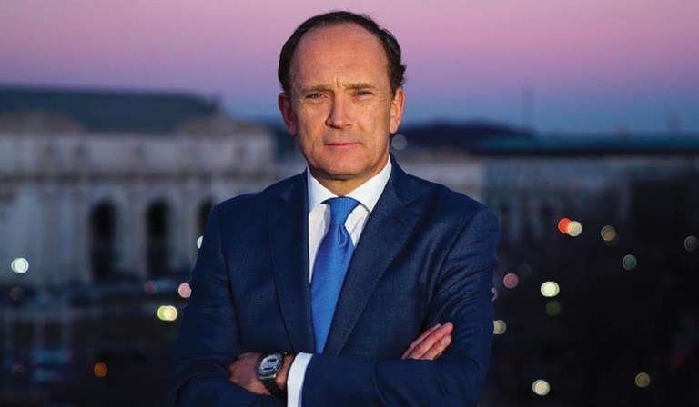 A white man with dark hair wears a navy suit and stands with his arms crossed against a purple sunset backdrop