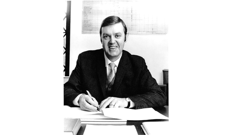 A black and white photo of a white man in a suit sitting at a desk and writing on a notepad