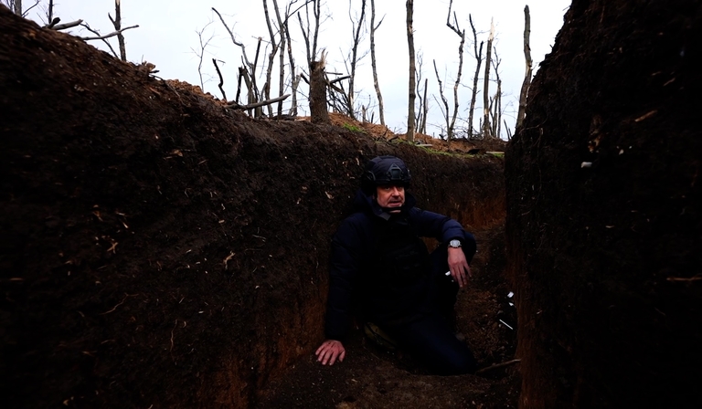 A news reporter sits in a trench in a body armour vest and helmet