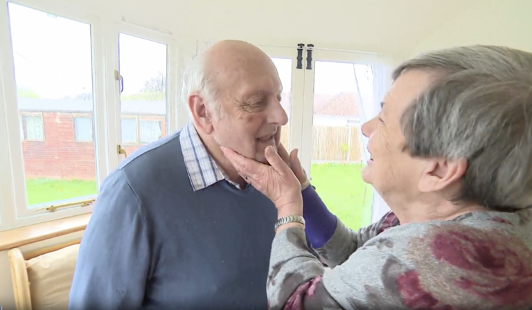 An elderly woman cradles an elderly man's chin in her hands